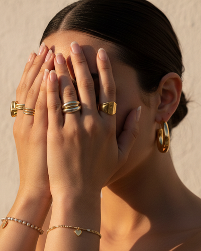 Candid, sunlit close-up of a Gen Z model's hands covering her face, with slicked-back dark hair. She is wearing a delicate beaded bracelet with a heart charm, several thin gold rings, and a prominent two-tone chunky ring. Her nails have a minimalist, barely-there polish. Large, sculptural gold earrings are visible, complementing the warm glow of the golden hour lighting against a textured, light background, evoking a playful and stylish mood.