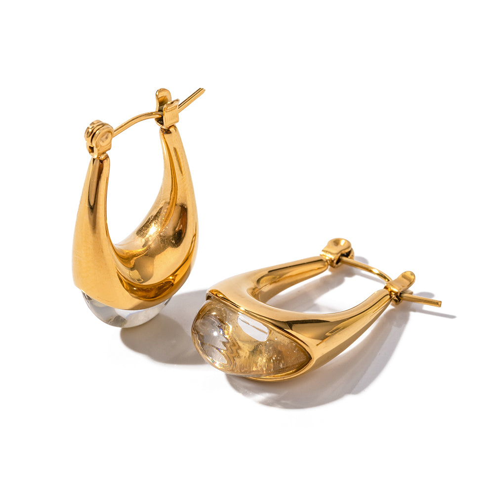 Gold tone huggie hoop earrings, 18K gold plated, with a rounded sculpted hoop shape and a glossy clear drop-like accent, hinged closure visible, no other stones visible.