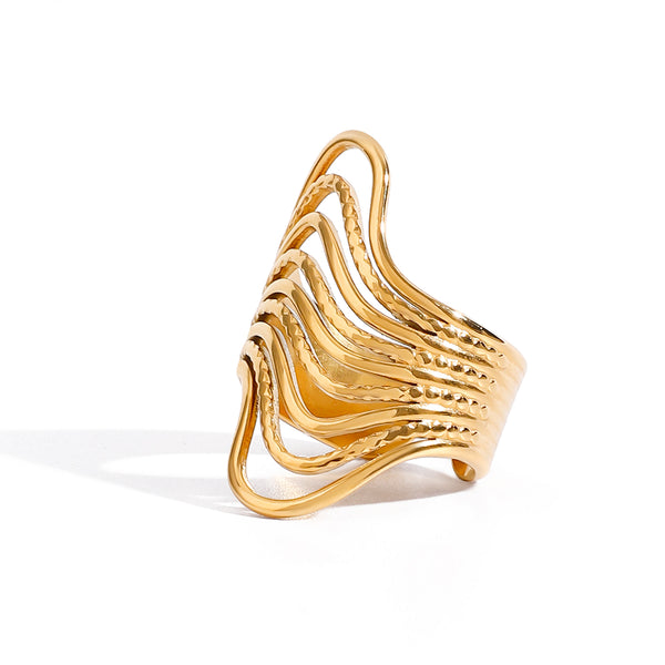 Gold tone statement ring, 18K gold plated, with a flowing wave silhouette, layered ridged bands, and lightly hammered texture, open cuff construction, no stones visible.